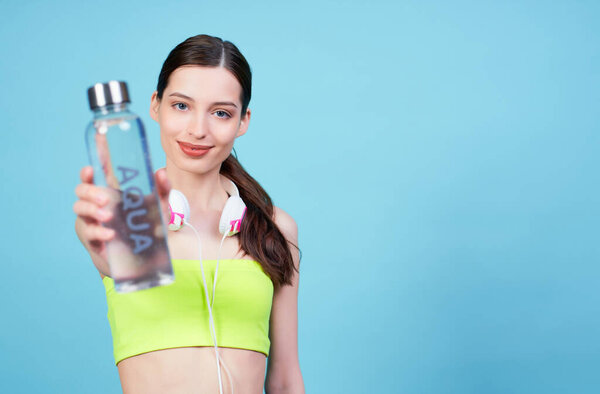 Charming athletic young Caucasian girl, in headphones and a light green top, posing on a blue background, holding a water bottle with the word aqua. Copy space.