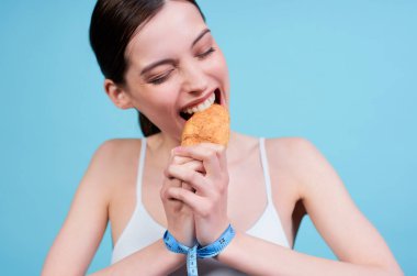 Yakın plan, beyaz bluzlu, kahverengi saçlı, güzel beyaz saçlı, meze bandıyla bağlanmış ve zevkle kruvasanı ısıran genç bir beyaz kadın. Diyet ve spor kavramı.