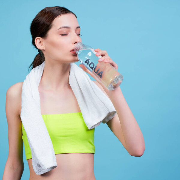 Close-up, athletic young slim white girl in a light green sports top, with a white towel on her neck, drinks water from a bottle after a workout, stands on a blue background.