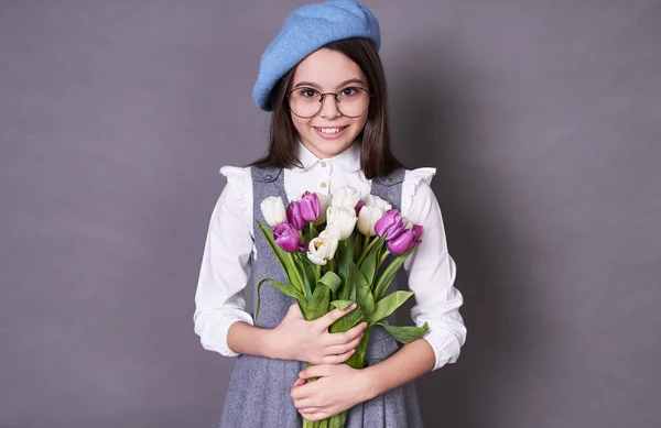 美しいかわいい若い感情的な女の子長い髪のブルネット スタイリッシュなブルーベレー帽で 彼女の手の中に花のチューリップのガラスでカメラを見て コピースペース 学校9月1 灰色の背景に — ストック写真