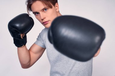 Yakışıklı kaslı atlet, mavi gözlü sarışın, gri tişörtlü, beyaz arka planda siyah boks eldivenli poz veren, kameraya bakan.