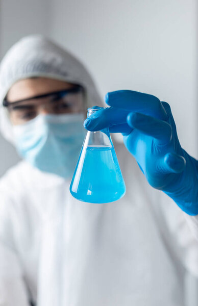 исследователь в голубых латексных перчатках и защитной одежде от коронавируса, смотрящий во фляжку с голубой жидкостью

