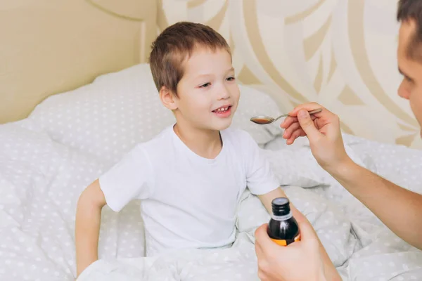 a blond-haired boy is sick, sitting in bed, he is given a drink of liquid medicine in a spoon