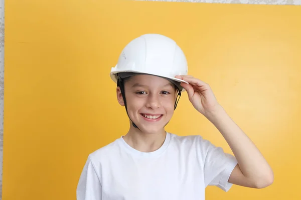 Caucasian boy with dark hair in a white t-shirt and a white protective construction safety helmet on a yellow background. Concept of labor protection and protection from external influences.