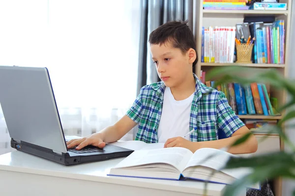 Distance learning online education. Schoolboy boy studies at home with laptop and does school homework. Training books and notebooks on table.