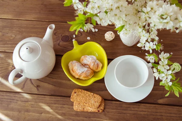 Baked croissants, cookies and a bouquet of white flowers in a vase on a dark wooden table, a white teapot and Cup. Setting the table for Breakfast and tea, good morning.