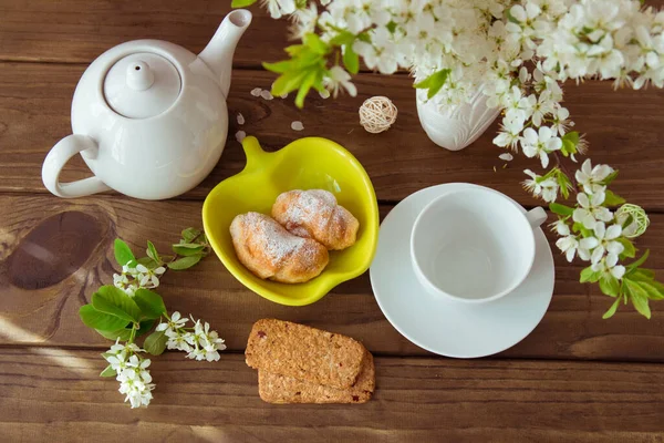 Baked croissants, cookies and a bouquet of white flowers in a vase on a dark wooden table, a white teapot and Cup. Setting the table for Breakfast and tea, good morning.