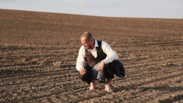 Porträt eines Mannes im klassischen Anzug barfuß auf dem Feld. Ein schöner bärtiger Mann sitzt auf einem Feld auf gepflügtem Land. ein Mann auf dem Feld berührt die Erde mit seiner Hand. — Stockvideo