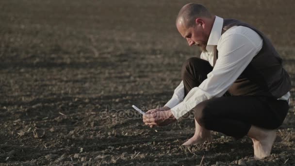 Un hombre agrónomo toma una foto de una plántula por teléfono. Retrato de un hombre barbudo en un campo con un teléfono móvil. Un hombre descalzo en el campo toma fotos de plantas . — Vídeos de Stock
