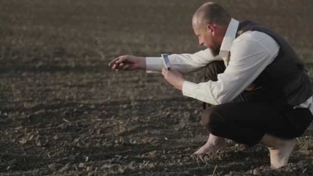 Un hombre agrónomo toma una foto de una plántula por teléfono. Retrato de un hombre barbudo en un campo con un teléfono móvil. Un hombre descalzo en el campo toma fotos de plantas . — Vídeos de Stock