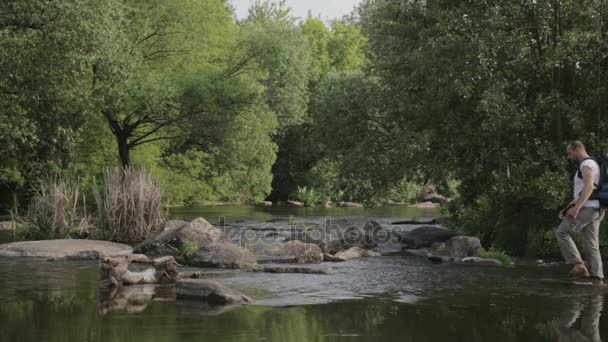 Guy bebaarde man reizen langs de rivier ford. Een toerist met een rugzak kruist de rivier. De bebaarde man kruist de rivier. — Stockvideo