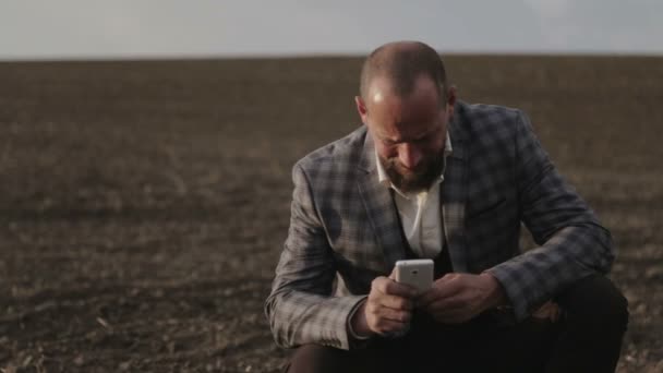 Un terrateniente barbudo está hablando en el campo por teléfono. Un hombre con traje habla emocionalmente en el móvil. Un hombre con barba en el campo con un teléfono . — Vídeos de Stock