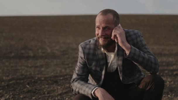 Un terrateniente barbudo está hablando en el campo por teléfono. Un hombre con traje habla emocionalmente en el móvil. Un hombre con barba en el campo con un teléfono . — Vídeos de Stock