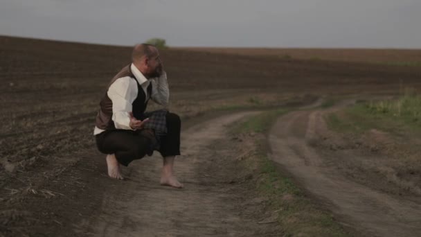 Un terrateniente barbudo está hablando en el campo por teléfono. Un hombre con traje habla emocionalmente en el móvil. Un hombre con barba en el campo con un teléfono . — Vídeos de Stock