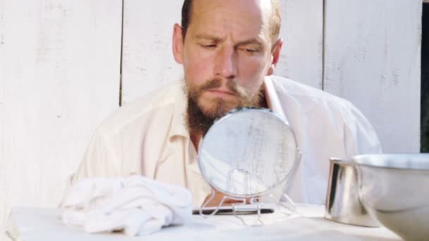 Een jongeman ziet er na zijn baard zittend aan een tafel. Portret van een bebaarde man bezig met zijn baard. Gezondheid, hygiëne, schoonheid. — Stockvideo