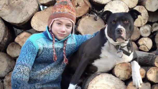 Il terrier dello Staffordshire lecca un ragazzo triste, sullo sfondo della legna da ardere. Ritratto di un bambino sullo sfondo della legna da ardere con un cane 4K video . — Video Stock