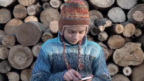 Um menino está jogando em um smartphone em uma aldeia perto da madeira. Adolescente na fazenda jogando no smartphone no jogo 4K vídeo . — Vídeo de Stock