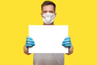 Hijyenik tıbbi maskeli ve elinde beyaz poster tutan mavi eldivenli bir adam. Model şablonu. Bilgi almak için reklam alanı. Coronavirus konsepti, COVID-19, kopyalama alanı.