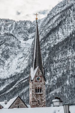 Kışın Hallstatt 'teki karlı dağın arka planına sahip taş kilise saat kulesi.
