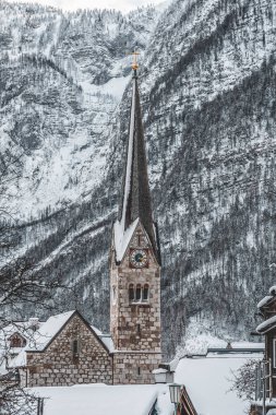 Kışın Hallstatt 'teki karlı dağın arka planına sahip taş kilise saat kulesi.