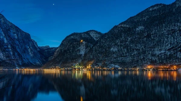 Austriacka ośnieżona wieś Hallstatt nad jeziorem otoczona śnieżnymi górami ze światłami zapalonymi po zachodzie słońca w nocy — Zdjęcie stockowe