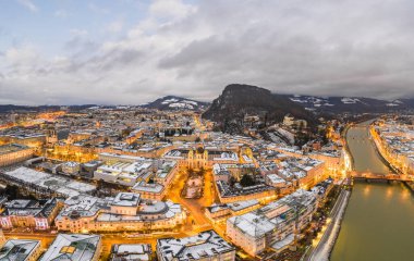 Salzburg 'daki Kutsal Üçlü Kilisesi' nin hava aracı görüntüsü kışın alacakaranlıkta karla kaplanırken Avusturya 'da şehir ışıkları açık.