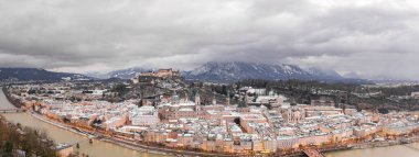 Panoramik insansız hava aracı, kış yağmurlarında sağanak bulutlar eşliğinde gün batımından sonra nehirden karlı Salzburg 'u görüntüledi.