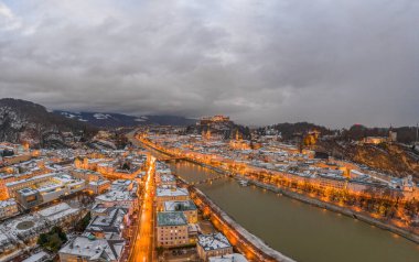 Panoramik insansız hava aracı, kış yağmurlarında sağanak bulutlar eşliğinde gün batımından sonra nehirden karlı Salzburg 'u görüntüledi.