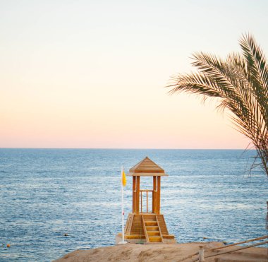 Güneş batarken tropik okyanusta cankurtaranlar için ahşap kule, gökyüzünde palmiye yaprakları.