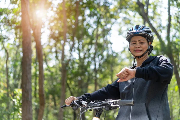 Sênior asiático mulher bicicleta no parque, com verificação de tempo ou ele — Fotografia de Stock