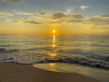 İnanılmaz güzel, muhteşem bir gün batımı, kumlu bir sahilde gün doğumu. Arkaplanda sakin bir deniz ve mavi-turuncu gökyüzü.