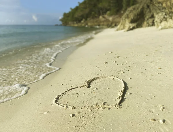 Kumlu sarı bir plaja yakın bir kalp çizilmiş. Denizin arka planında beyaz deniz köpüğü, mavi gökyüzü ve ağaçlı kayalık kıyı..
