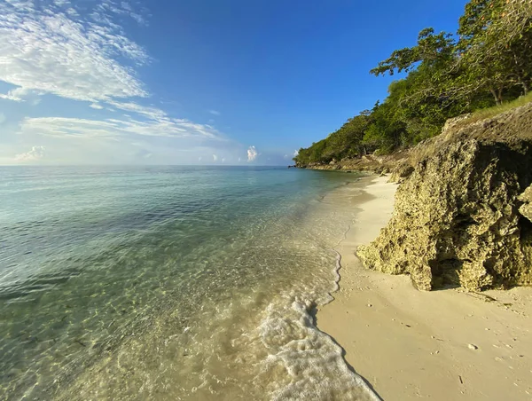 Beyaz kumlu bir sahil ve turkuaz şeffaf bir deniz manzarası. Yeşil ormanlı kayalık sahil.