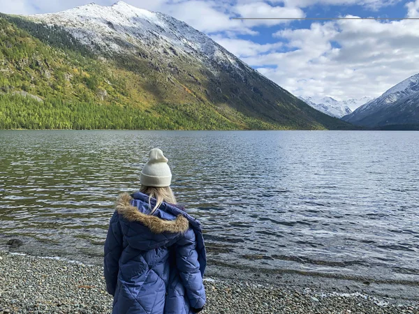 Şapkalı ve ceketli sarışın kız, saydam suyla gölün arka planındaki çakıl taşı plajında duruyor. Yüzü olmayan kişi kar kaplı güzel dağlara bakıyor..