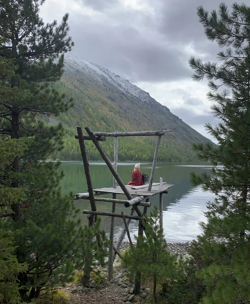 Arkadan gelen sarışın kız eski bir iskelede ekose bir battaniyenin içinde oturur, termostan gelen bir su ile kendini ısıtır. Yeşil şeffaf göl manzarası, kozalaklı orman, karla kaplı dağlar.