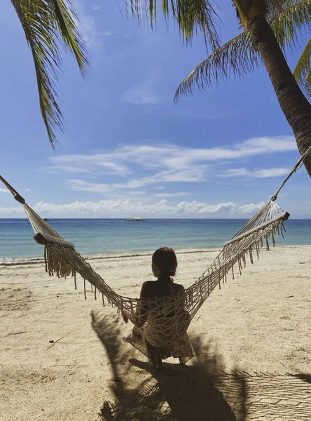 Una ragazza senza faccia è seduta su un'amaca su una spiaggia di sabbia bianca. Una donna dal retro guarda il mare turchese e il cielo blu. Una persona si rilassa in un bellissimo resort — Foto Stock