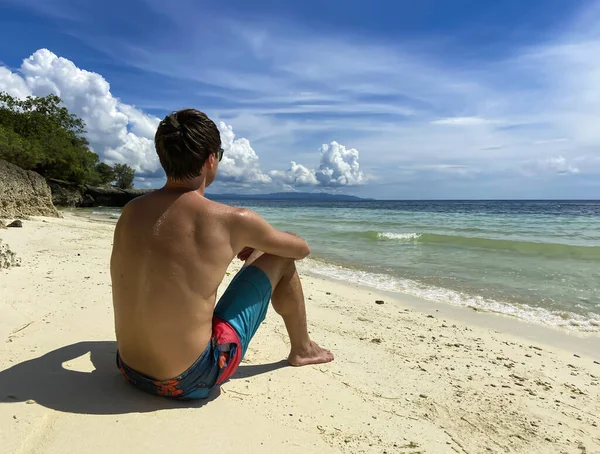 Arkadan gözlüklü adam beyaz kumlu sahilde oturuyor ve turkuaz denize bakıyor. Yüzü olmayan adam güzel sahilde dinleniyor.