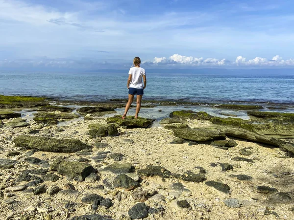 Sarışın kız sırtı taş levhalar ve kumla sahilde duruyor. Gökyüzü ve bulutların arkasında mavi deniz ve kadının güzel manzarası.
