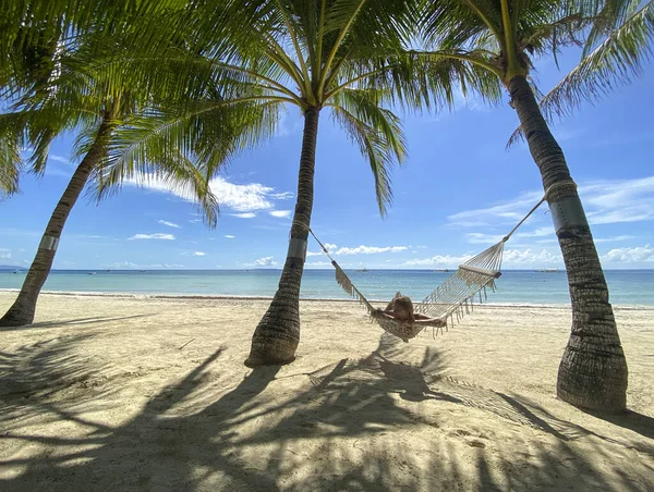 Girl resting and swinging on hammock in paradise resort.Beautiful sandy white beach with turquoise sea in grove with palm trees. Shadows on sand — Stock Photo, Image
