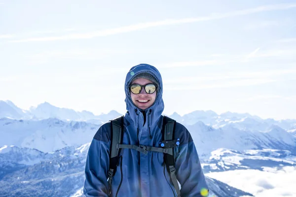 Montañista mochilero sonriendo en la cumbre contra las capas nevadas azules de la montaña en invierno. cielo azul, soleado Fotos De Stock