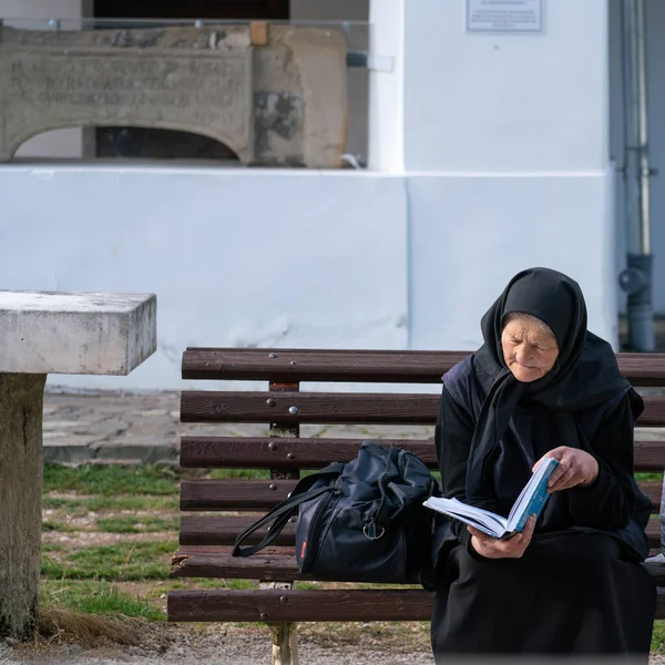 Sinaia, Romanya - 16 Kasım 2019: Ortodoks Katolik Hristiyanlığını uygulayan yaşlı dindar bir kadın, Sinaia Manastırı 'nın önündeki bir bankta oturmuş, bir kitap okuyor.