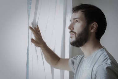 Endişeli bir yüzü olan sakallı genç adam, umut dolu bir bakışla evinin penceresinden bakıyor. Coronavirus için kendi kampanyanda kal..