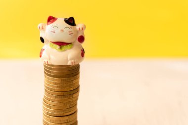 Maneki neko ya da bozuk para yığınına konmuş işaret eden kedi. Şansın, bolluğun ve ekonomik başarının kavramsal sembolü. Sarı arkaplan.