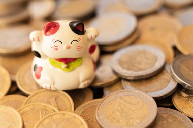 Maneki neko ya da işaret eden kedi dağınık para yığınının üzerine yerleştirilmiş. Şansın, bolluğun ve ekonomik başarının kavramsal sembolü.