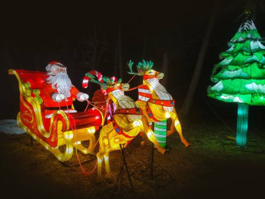 Noel Baba 'nın aydınlanmış figürü, kış mevsiminde Noel festivalinde gece vakti. Bir Noel geyiği ve kızağını resmeden ampullerden oluşan bir kompozisyon..