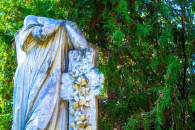 Mavi gökyüzüne ve mezar taşına karşı kolları çaprazlanmış, haç arkaplanlı diz çökmüş bir meleğin başı kesilmiş heykeli.