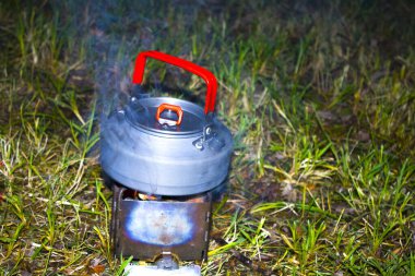 Kahve demlerken ateş çatırdıyor. Açık havada pişirmek için katlanmış odun sobası. Doğanın arka planında yakıt olarak ince dallar kullanılıyor. Kahve demliği ve ateşte güneşlenen kahve.