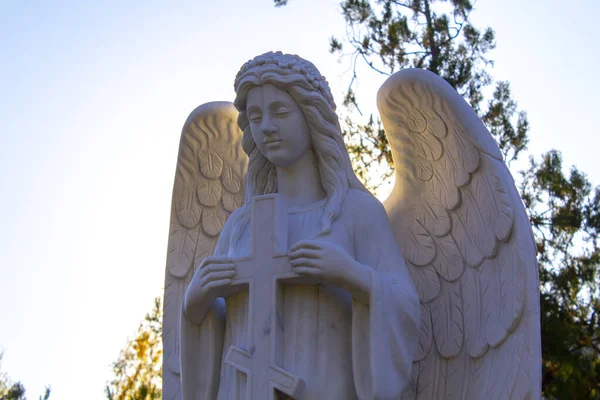 statue of kneeling angel with arms crossed against blue sky and cemetery headstone with crucifix background