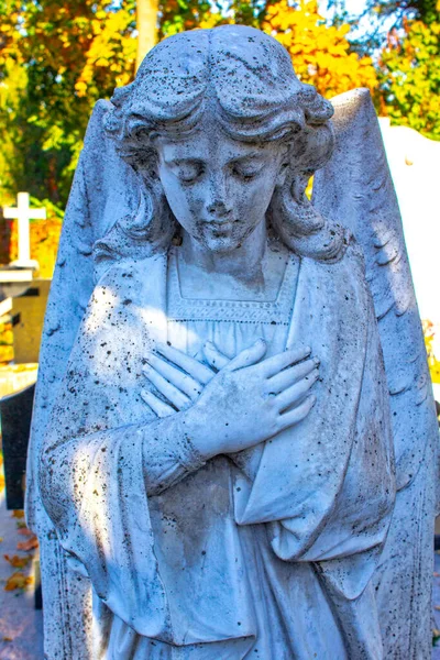 statue of kneeling angel with arms crossed against blue sky and cemetery headstone with crucifix background