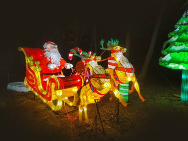 Noel Baba 'nın aydınlanmış figürü, kış mevsiminde Noel festivalinde gece vakti. Bir Noel geyiği ve kızağını resmeden ampullerden oluşan bir kompozisyon..